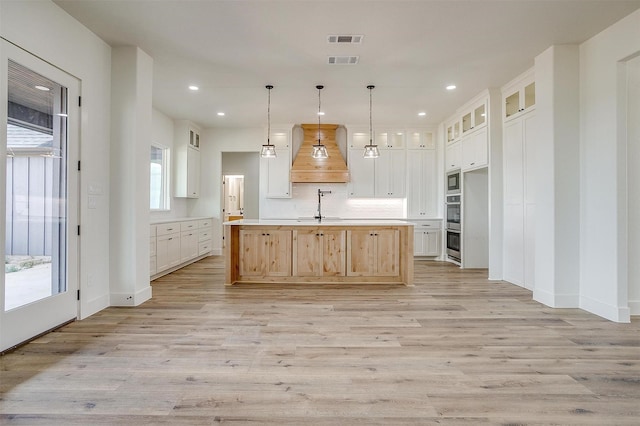 kitchen featuring premium range hood, a large island with sink, white cabinets, light wood-type flooring, and decorative light fixtures