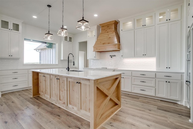 kitchen featuring custom exhaust hood, light countertops, glass insert cabinets, a sink, and an island with sink