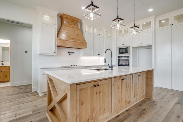 kitchen featuring custom range hood, glass insert cabinets, white cabinetry, a sink, and an island with sink