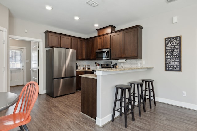 kitchen with light stone countertops, stainless steel appliances, wood-type flooring, kitchen peninsula, and decorative backsplash