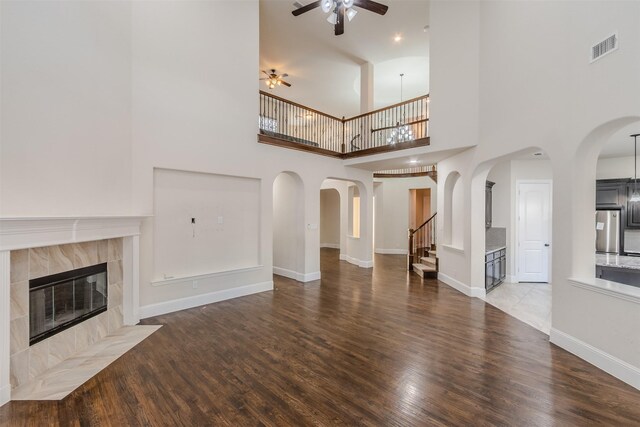 unfurnished living room with a high ceiling, ceiling fan with notable chandelier, a tiled fireplace, and wood-type flooring