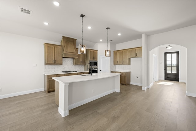 kitchen with light wood-type flooring, custom range hood, gas stovetop, stainless steel microwave, and an island with sink