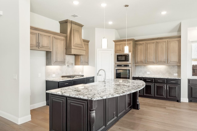 kitchen featuring premium range hood, a kitchen island with sink, stainless steel appliances, light stone counters, and light hardwood / wood-style floors