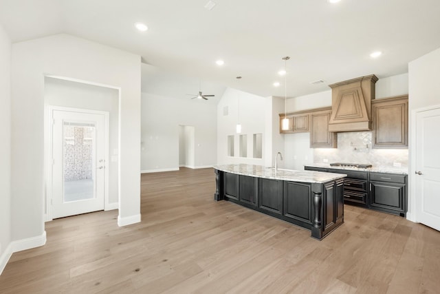 kitchen featuring stainless steel gas stovetop, decorative backsplash, custom exhaust hood, a center island with sink, and light wood-type flooring