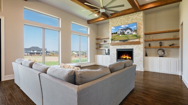 living room featuring beamed ceiling, a stone fireplace, built in shelves, dark wood-type flooring, and ceiling fan