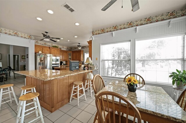 kitchen with light tile patterned floors, appliances with stainless steel finishes, light stone countertops, ceiling fan, and a breakfast bar