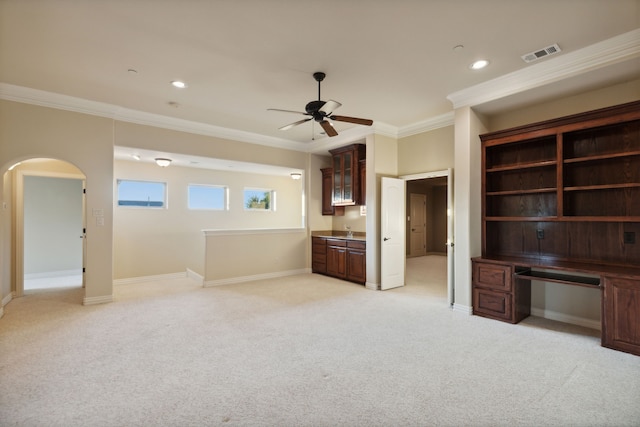 unfurnished living room with light carpet, crown molding, built in desk, and ceiling fan