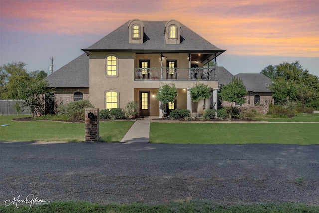 french country style house featuring a balcony, stucco siding, a shingled roof, and a yard