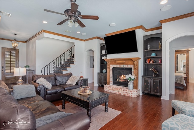 living room with a brick fireplace, dark wood-type flooring, built in features, ornamental molding, and ceiling fan with notable chandelier