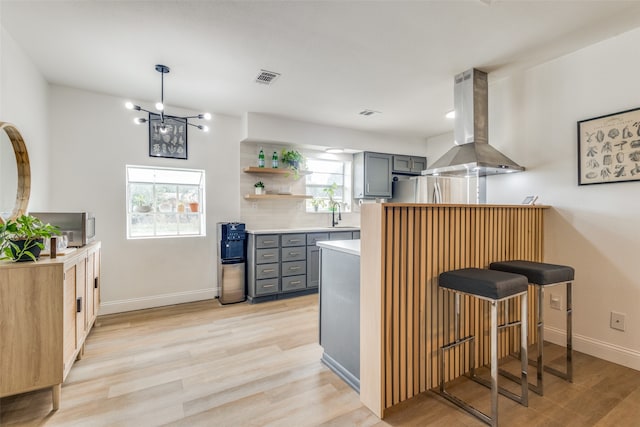 kitchen with light hardwood / wood-style floors, a notable chandelier, gray cabinets, wall chimney exhaust hood, and kitchen peninsula