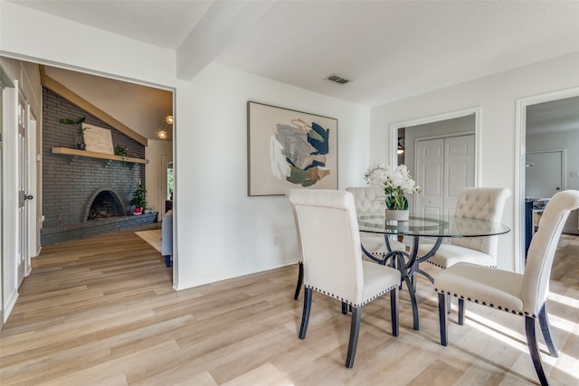 dining room featuring a textured ceiling, light hardwood / wood-style flooring, a brick fireplace, brick wall, and vaulted ceiling with beams