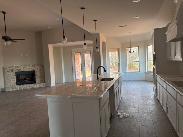 kitchen featuring a center island with sink, a stone fireplace, sink, decorative light fixtures, and light stone counters