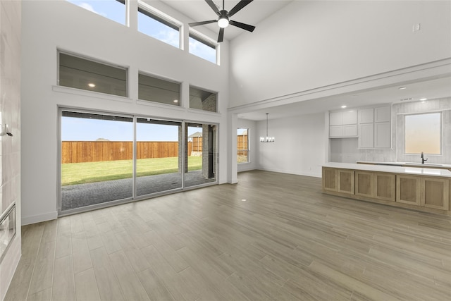 unfurnished living room featuring light wood-type flooring, a wealth of natural light, ceiling fan with notable chandelier, and a towering ceiling