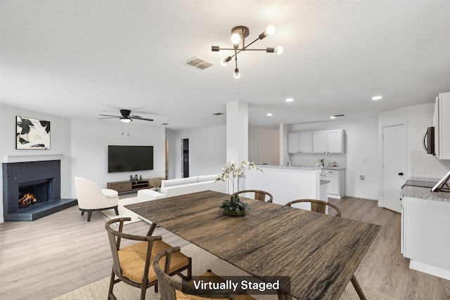 dining area with light wood-type flooring, ceiling fan with notable chandelier, a fireplace, and a textured ceiling