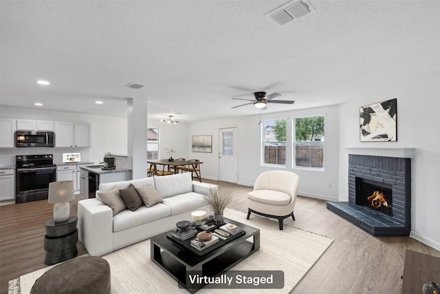 living room featuring a brick fireplace, light wood-type flooring, ceiling fan, and a textured ceiling