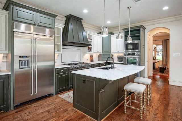 kitchen with crown molding, built in appliances, light stone countertops, dark wood-type flooring, and a center island with sink