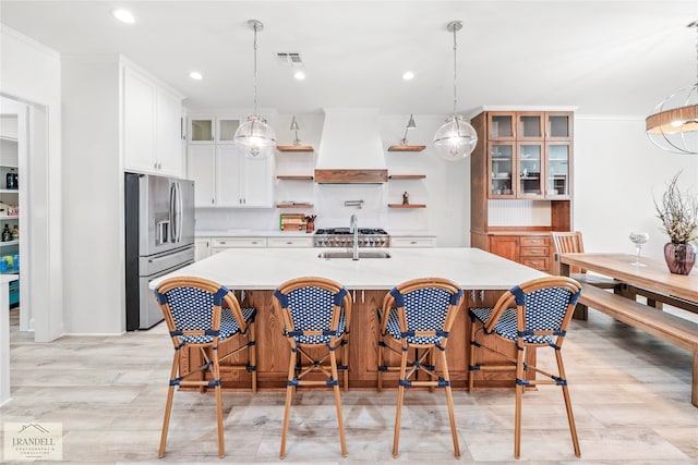 kitchen featuring white cabinets, premium range hood, stainless steel refrigerator with ice dispenser, pendant lighting, and a kitchen island with sink