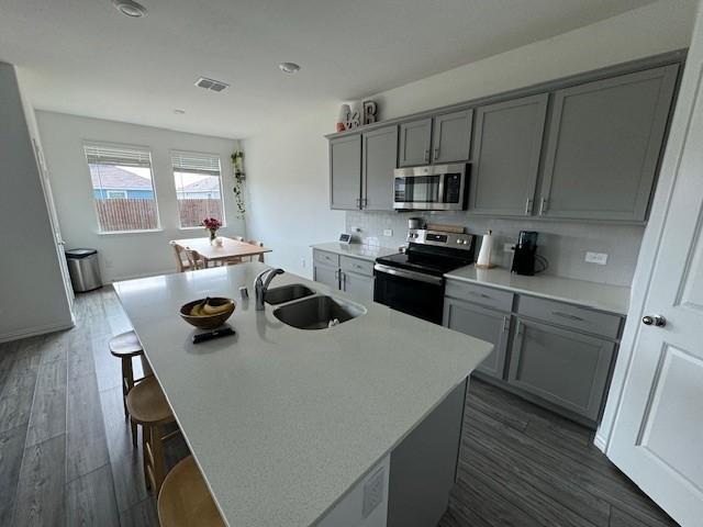 kitchen with gray cabinets, sink, dark wood-type flooring, appliances with stainless steel finishes, and a center island with sink