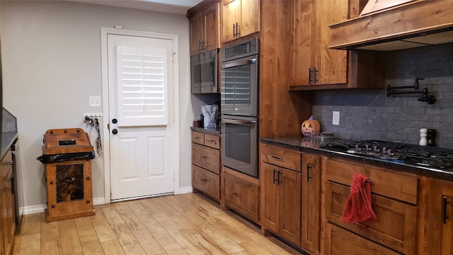 kitchen featuring premium range hood, backsplash, stainless steel appliances, dark stone counters, and light wood-type flooring