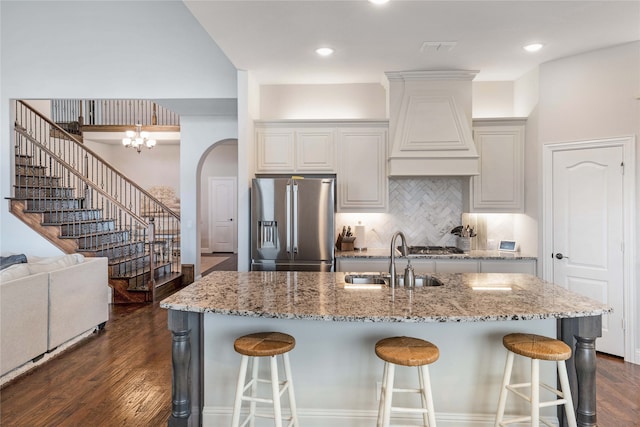 kitchen with a kitchen island with sink, stainless steel appliances, premium range hood, sink, and dark wood-type flooring