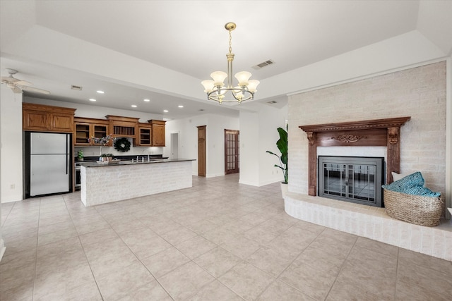 unfurnished living room with light tile patterned floors, a brick fireplace, ceiling fan with notable chandelier, sink, and a tray ceiling