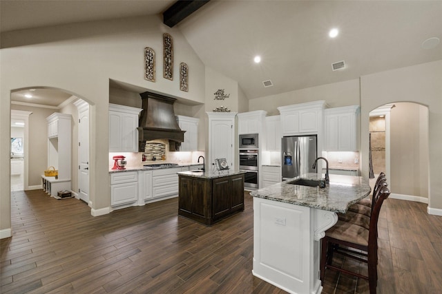 kitchen with arched walkways, stainless steel appliances, custom range hood, a kitchen island with sink, and a sink