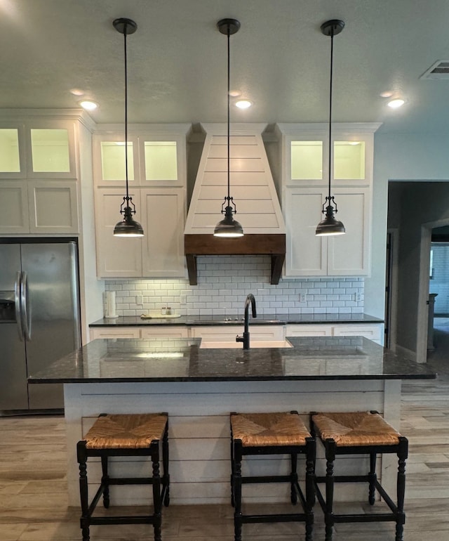 kitchen with custom range hood, light hardwood / wood-style floors, white cabinetry, stainless steel refrigerator with ice dispenser, and a kitchen island with sink