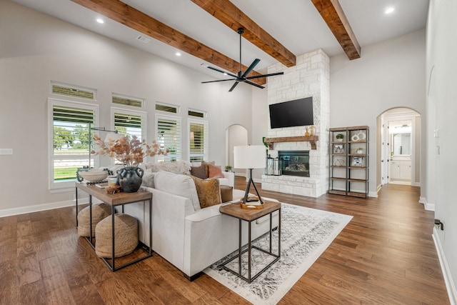 living room with beamed ceiling, wood-type flooring, a stone fireplace, ceiling fan, and a towering ceiling