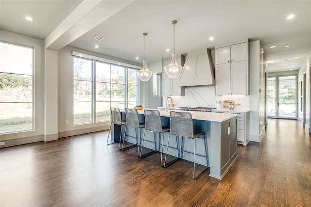 kitchen with tasteful backsplash, custom exhaust hood, decorative light fixtures, a kitchen island with sink, and dark hardwood / wood-style floors