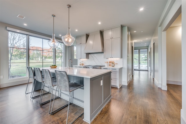 kitchen with backsplash, pendant lighting, wood-type flooring, custom exhaust hood, and a center island with sink