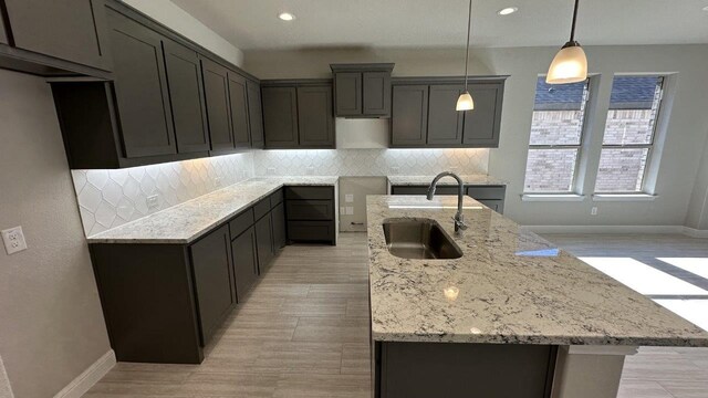 kitchen featuring decorative backsplash, sink, hanging light fixtures, a kitchen island with sink, and light stone countertops