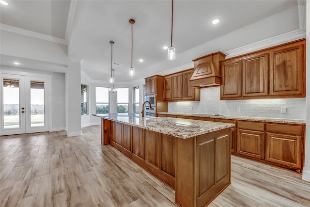 kitchen with light stone countertops, premium range hood, decorative light fixtures, light hardwood / wood-style floors, and an island with sink