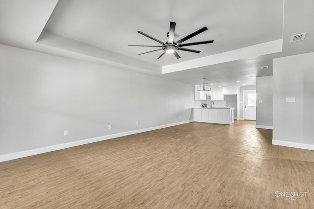 unfurnished living room featuring light hardwood / wood-style floors, sink, a tray ceiling, and ceiling fan with notable chandelier