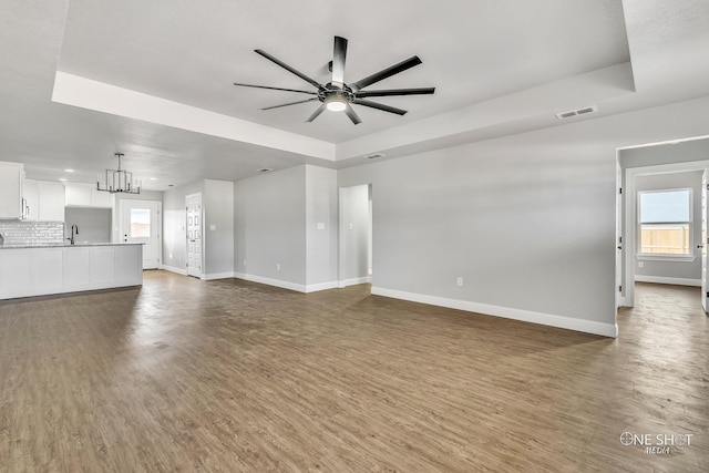 unfurnished living room featuring ceiling fan with notable chandelier, a tray ceiling, and dark hardwood / wood-style floors