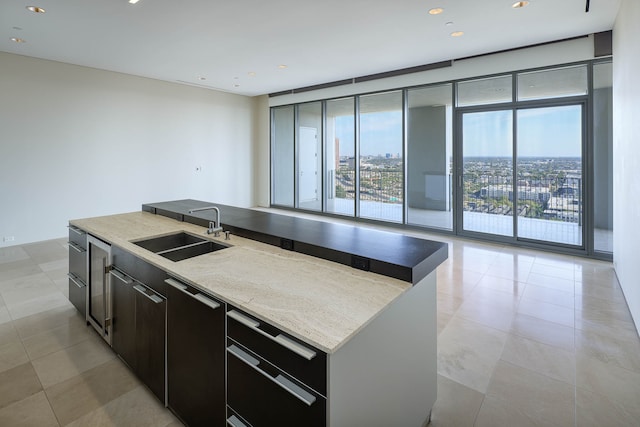 kitchen with an island with sink, sink, light tile patterned flooring, expansive windows, and light stone counters