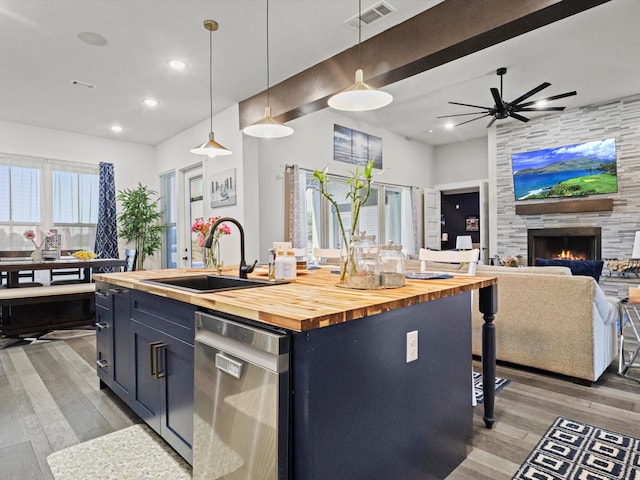 kitchen featuring hanging light fixtures, a fireplace, a center island with sink, stainless steel dishwasher, and wood counters