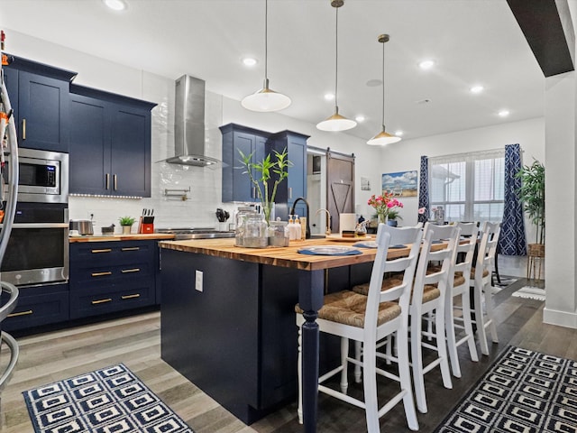 kitchen with a kitchen island with sink, butcher block counters, a kitchen bar, wall chimney range hood, and stainless steel appliances