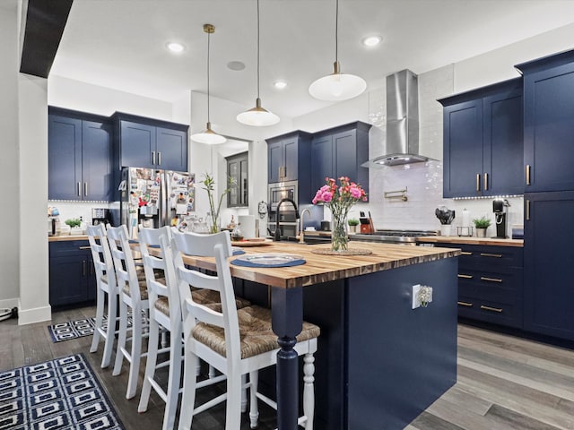 kitchen with wall chimney exhaust hood, decorative light fixtures, a kitchen island with sink, and wooden counters