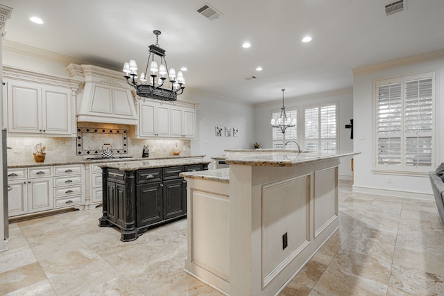 kitchen with light stone countertops, crown molding, a center island with sink, decorative light fixtures, and backsplash