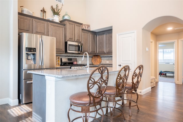 kitchen featuring light stone counters, dark hardwood / wood-style floors, stainless steel appliances, backsplash, and a center island with sink