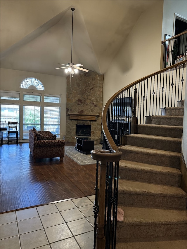 staircase with ceiling fan, a stone fireplace, high vaulted ceiling, and hardwood / wood-style flooring