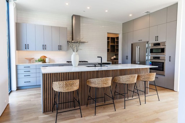 kitchen with appliances with stainless steel finishes, a breakfast bar, an island with sink, wall chimney exhaust hood, and gray cabinets