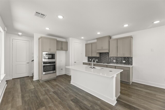 kitchen with appliances with stainless steel finishes, a center island with sink, gray cabinets, and dark wood-type flooring