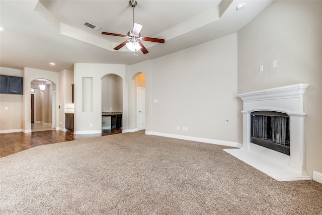 unfurnished living room featuring a raised ceiling, dark colored carpet, and ceiling fan