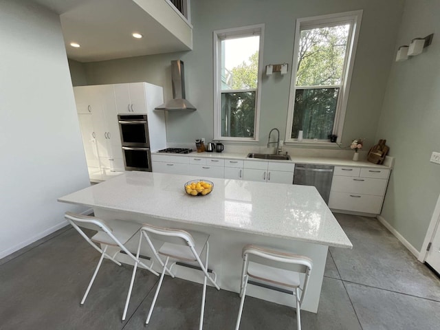 kitchen with white cabinetry, wall chimney exhaust hood, stainless steel appliances, a kitchen bar, and sink