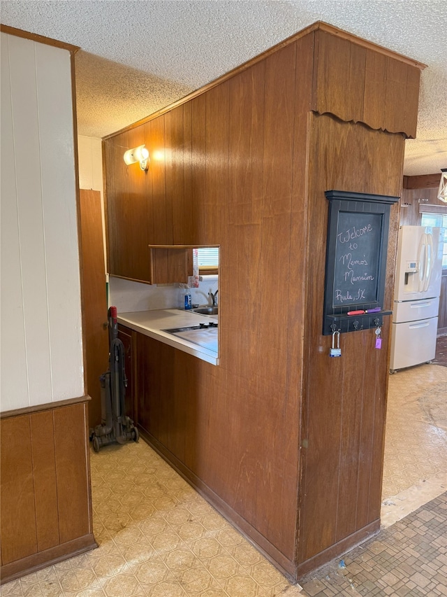 kitchen with a textured ceiling, light floors, wood walls, white fridge with ice dispenser, and brown cabinets