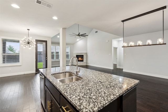 kitchen featuring ceiling fan with notable chandelier, a stone fireplace, decorative light fixtures, a kitchen island with sink, and sink