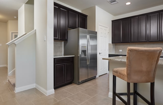 kitchen featuring dark brown cabinets, light stone counters, tasteful backsplash, light tile patterned floors, and stainless steel fridge