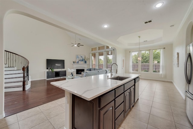 kitchen with light tile patterned floors, an island with sink, a raised ceiling, dark brown cabinets, and sink