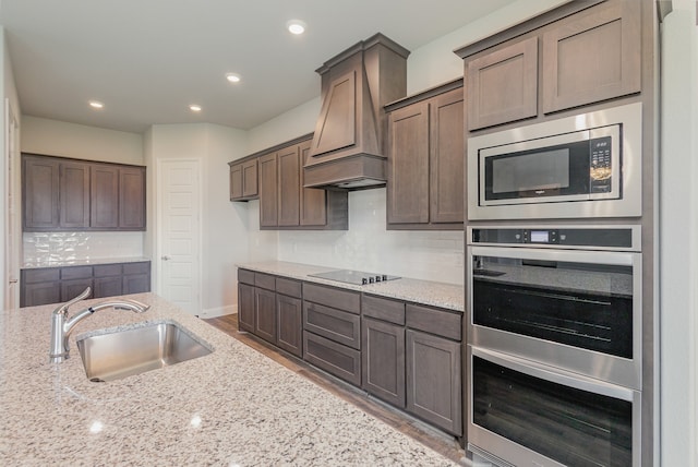 kitchen with light stone counters, custom exhaust hood, sink, dark brown cabinets, and stainless steel appliances